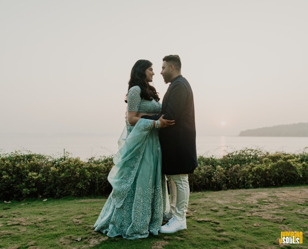 Couple embracing and smiling at each other near the coast at sunset; woman wears a teal traditional dress, man wears a dark blazer and light pants.