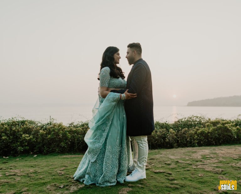 Couple embracing and smiling at each other near the coast at sunset; woman wears a teal traditional dress, man wears a dark blazer and light pants.