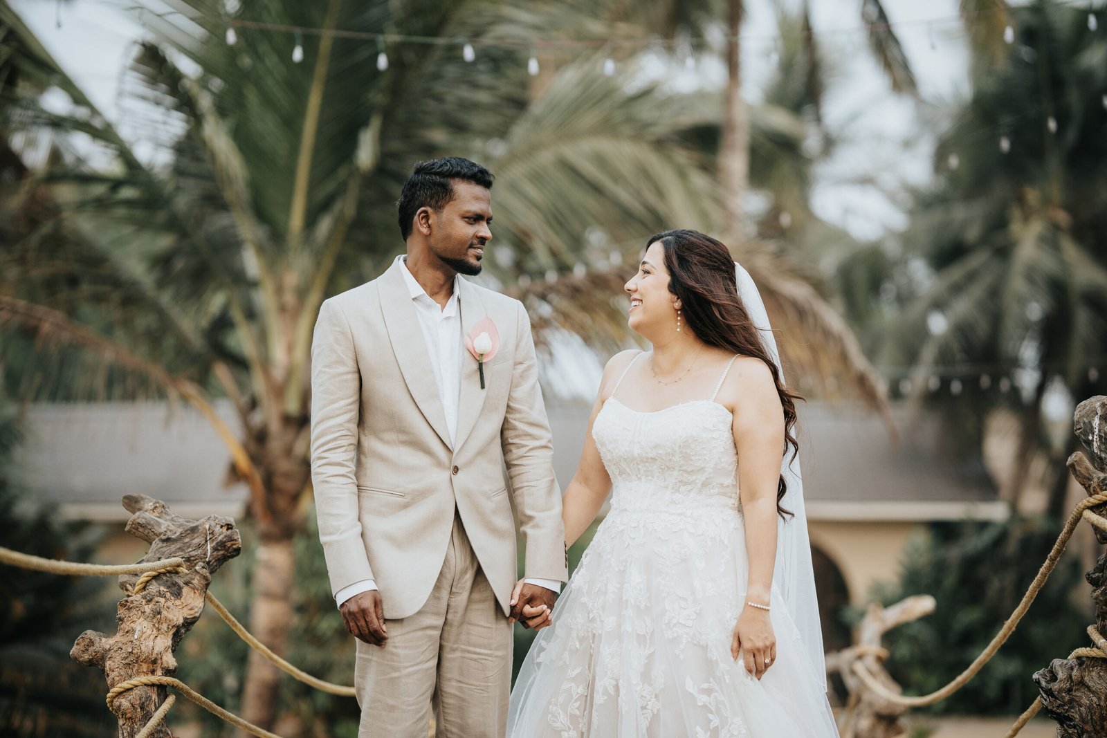 Bride in a white lace gown and groom in a light beige suit stand arm in arm outdoors in a tropical garden with palm trees; she holds a pink-and-white bouquet.
