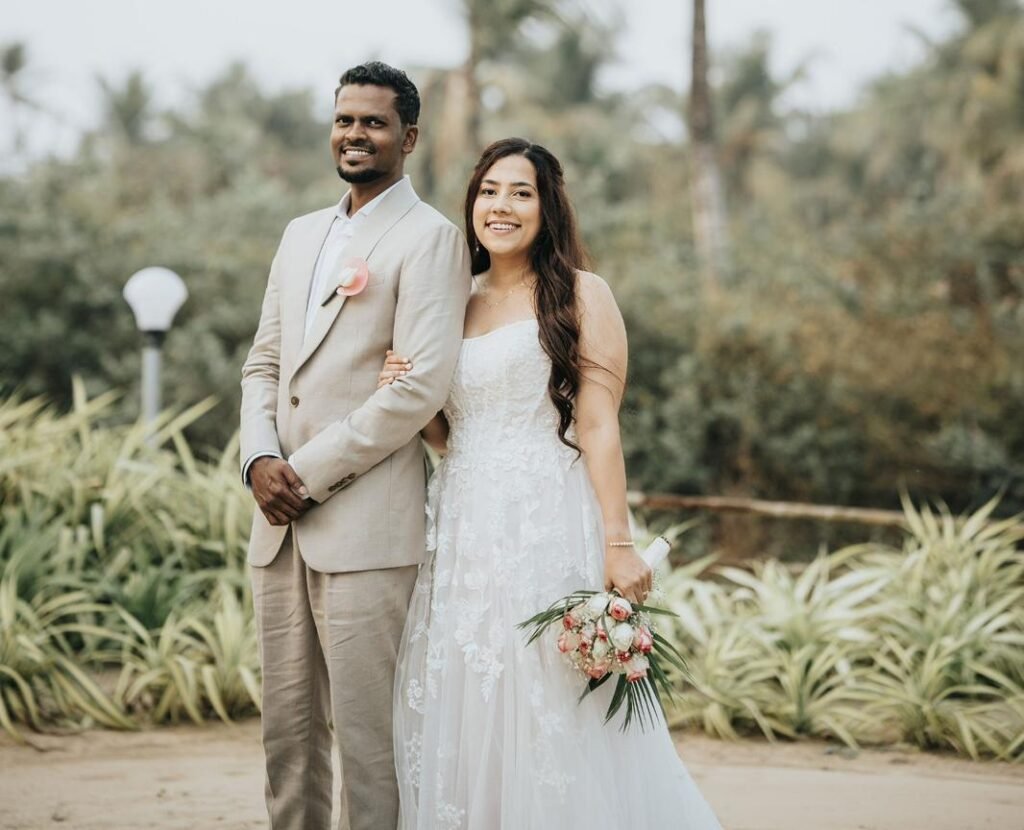 Bride in a white lace gown and groom in a light beige suit stand arm in arm outdoors in a tropical garden with palm trees; she holds a pink-and-white bouquet.
