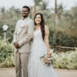 Bride in a white lace gown and groom in a light beige suit stand arm in arm outdoors in a tropical garden with palm trees; she holds a pink-and-white bouquet.