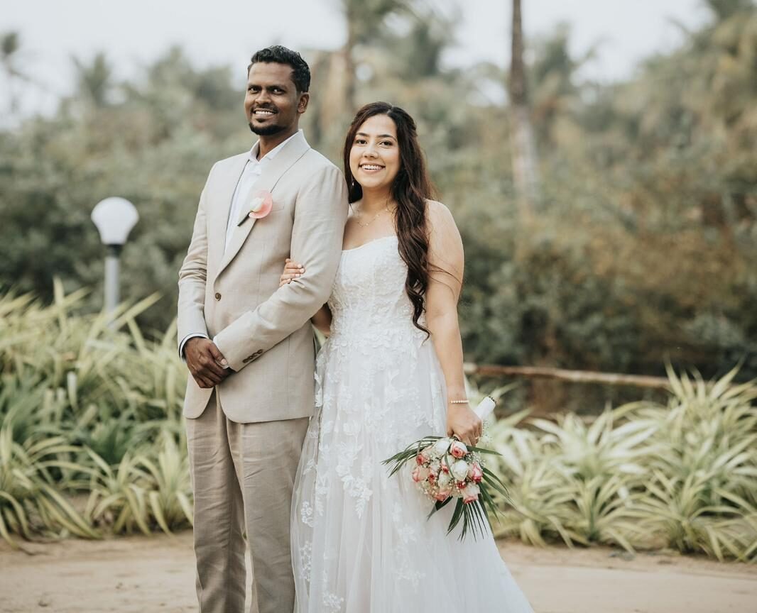 Bride in a white lace gown and groom in a light beige suit stand arm in arm outdoors in a tropical garden with palm trees; she holds a pink-and-white bouquet.