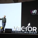 Speaker on a stage at a Vector Consulting Group event, pointing to the screen with a large projection behind him.