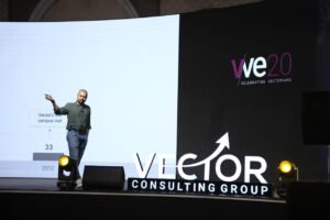 Speaker on a stage at a Vector Consulting Group event, pointing to the screen with a large projection behind him.