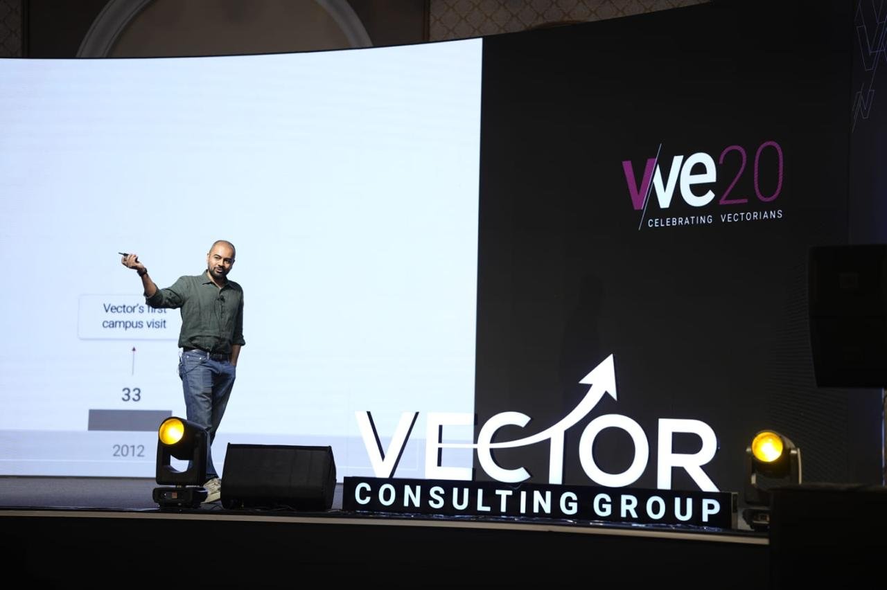 Speaker on a stage at a Vector Consulting Group event, pointing to the screen with a large projection behind him.