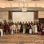 Large group portrait of 40+ people in formal attire posing in a hotel banquet hall with a projection screen behind them.