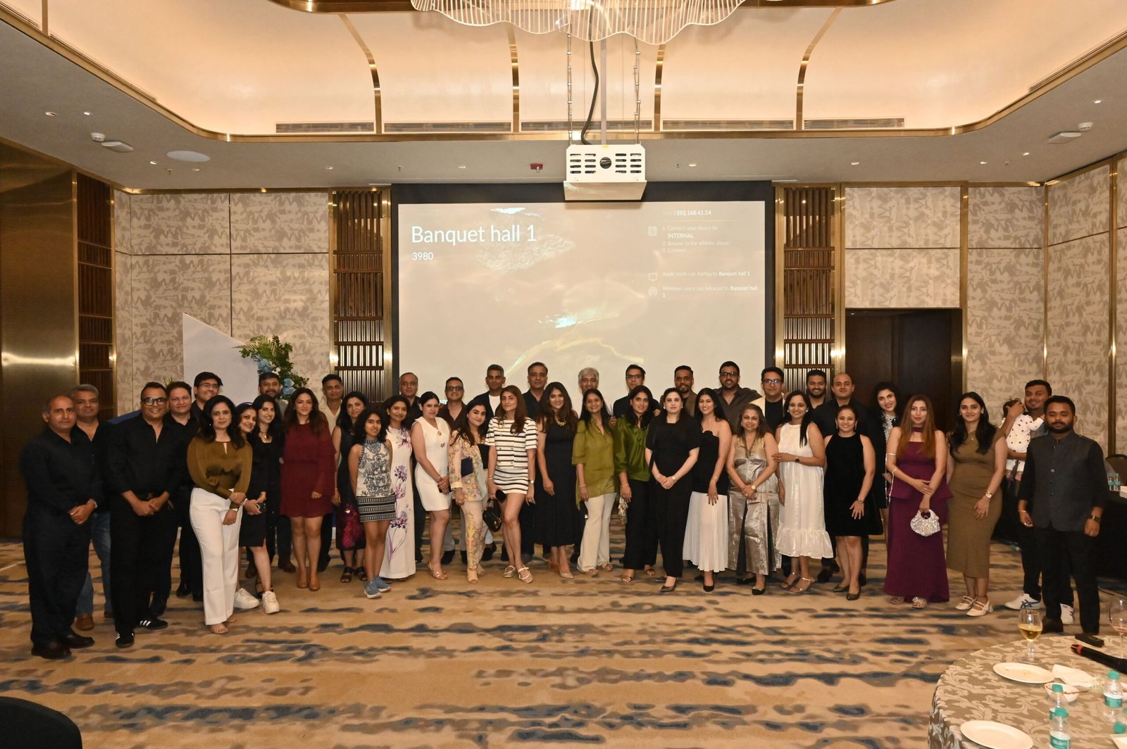 Large group portrait of 40+ people in formal attire posing in a hotel banquet hall with a projection screen behind them.