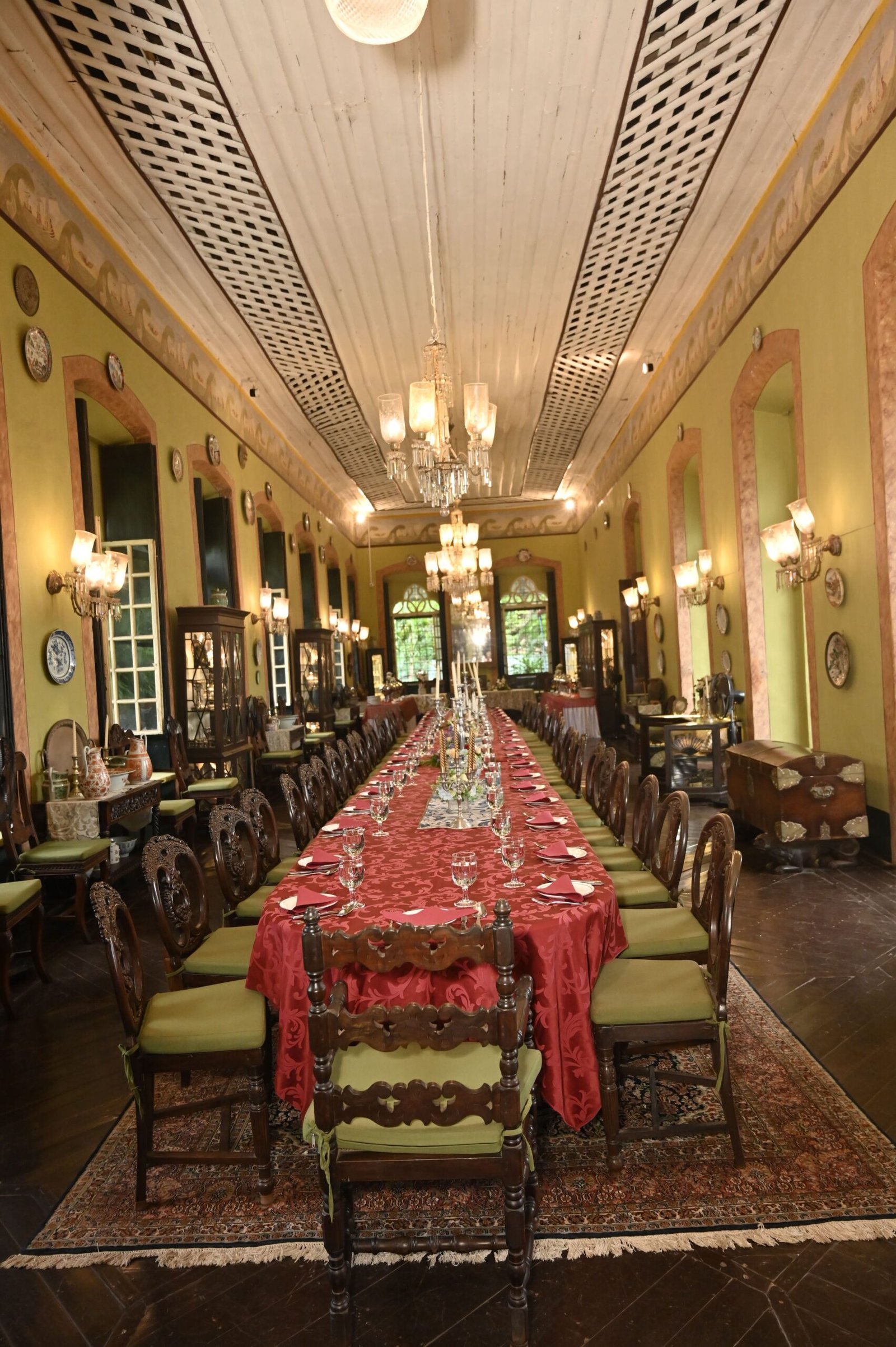 Long formal dining hall with a red table runner, ornate wooden chairs, and green walls adorned with plates and mirrors; chandeliers hang from a high, decorative ceiling.