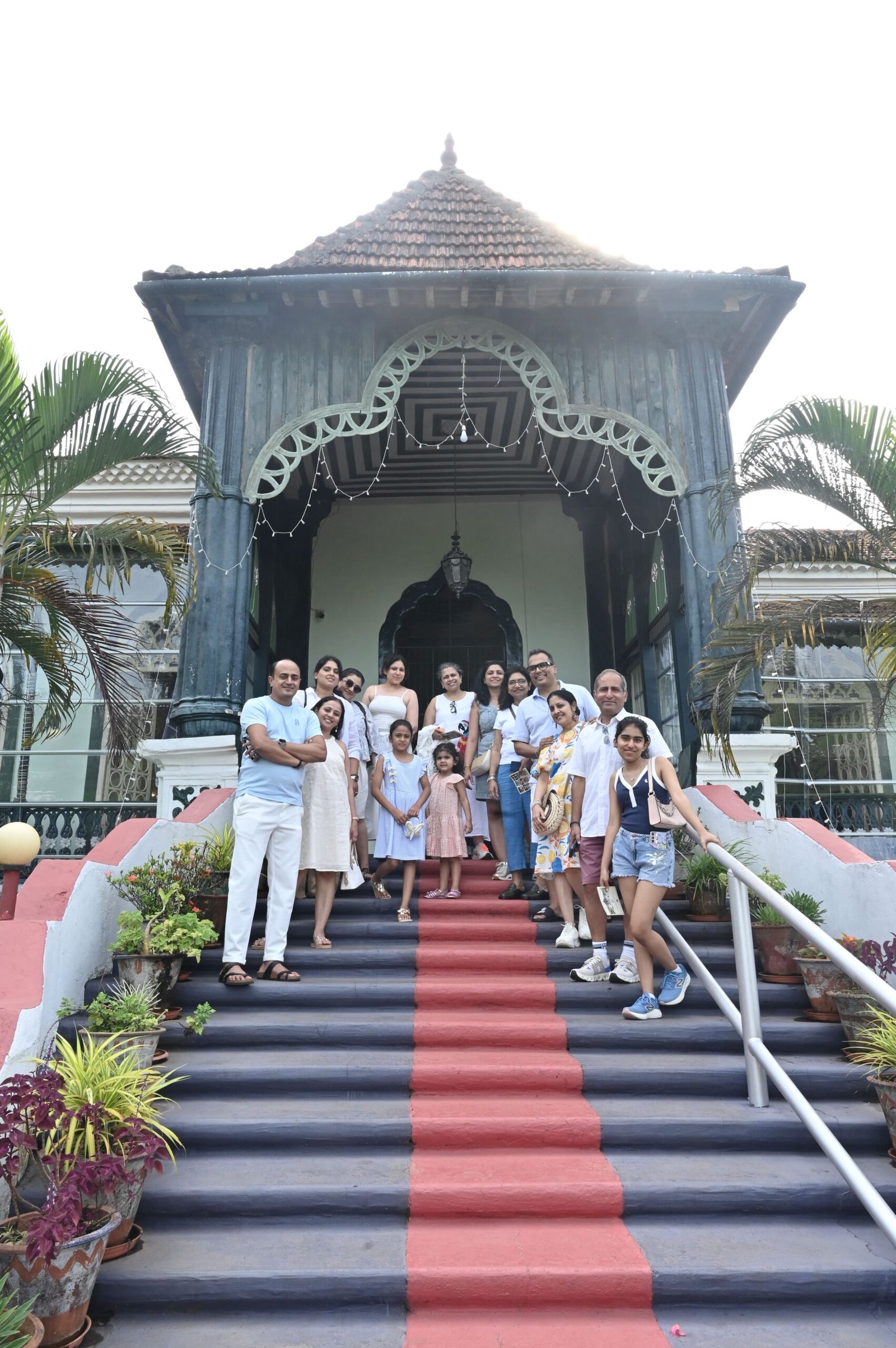 Group of people posing on a striped red-carpeted staircase in front of a blue wooden pavilion with a tiled roof.