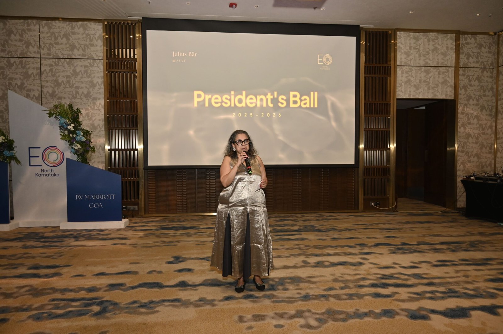 A woman in a metallic gown speaks into a microphone on a stage with a large screen reading "President's Ball 2025-2026" behind her.