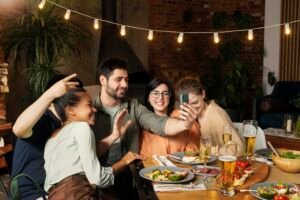 Group of friends taking a selfie at a dinner table, with plates and drinks in front of them in a cozy restaurant setting.