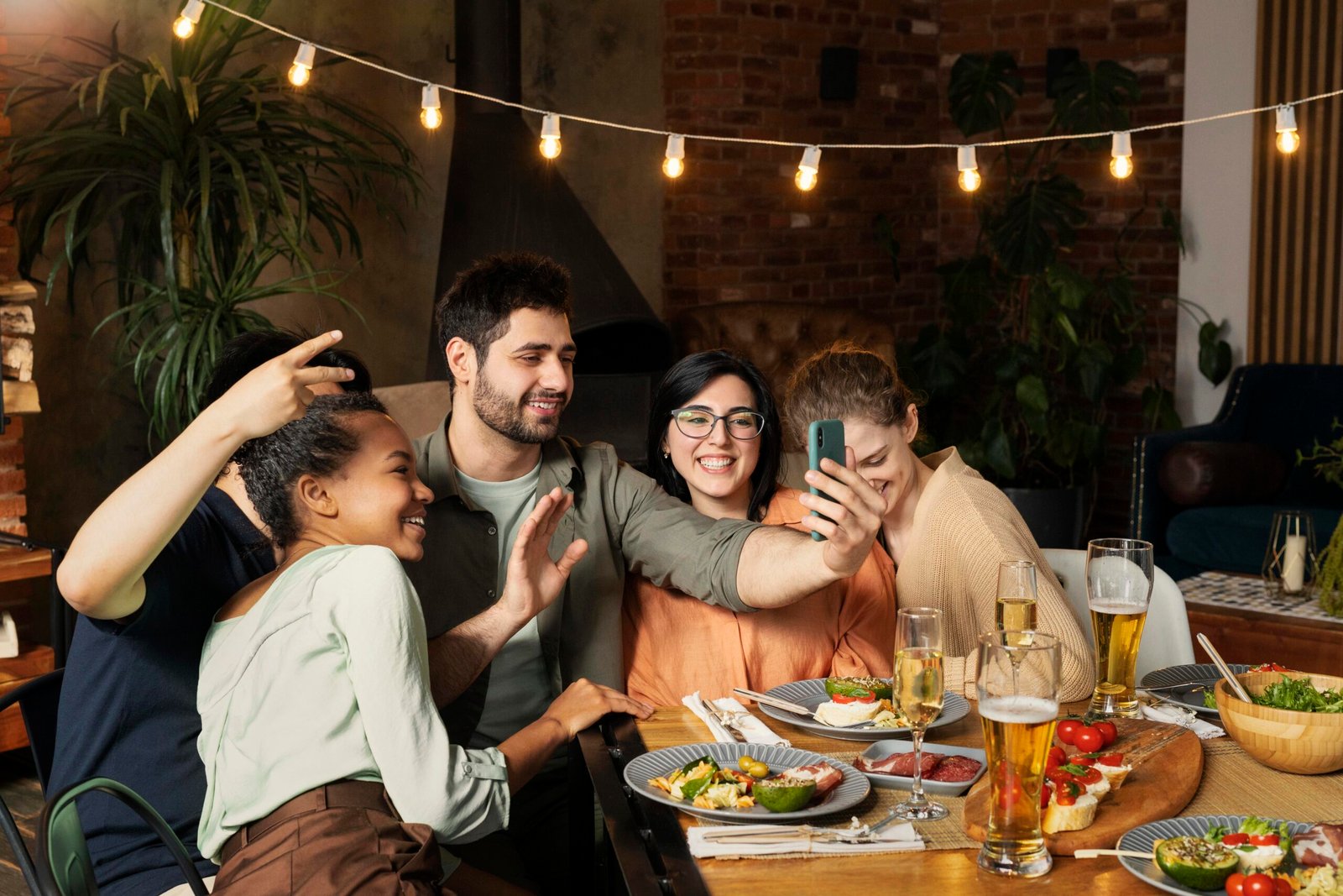 Group of friends taking a selfie at a dinner table, with plates and drinks in front of them in a cozy restaurant setting.