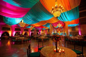 photorealistic-wedding-venue-with-intricate-decor-ornaments - XINANE Banquet hall decorated with vibrant turquoise, pink, orange drapes and gilded chandeliers above round glittering tables.